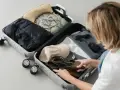 Kobieta pakuje do walizki: czarną kurtkę, zielony biustonosz, beżową torebkę, czapkę, buty, okulary i perfumy. Gotowa na podróż, pakuje swój enter air bagaż.