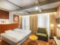 Przytulny pokój w hotelu Qubus Wrocław z dwoma łóżkami, zielonym fotelem i stolikiem. Widoczne betonowe belki na suficie.