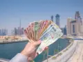 Dłoń trzymająca wachlarz banknotów dirhamów ZEA na tle panoramy Dubaju.
