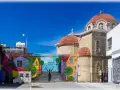 Kolorowy mural z domkami i rowerzystą zdobi ścianę w Pafos, stare miasto. Obok wznosi się kamienna cerkiew z pomarańczowymi kopułami.