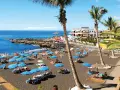 Czarna plaża Playa de la Arena na Teneryfie z turystami, niebieskimi parasolami i palmami.