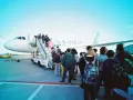 Pasażerowie wsiadają do samolotu. W tle widać żółtą taksówkę i personel lotniska. Zapalniczka w samolocie jest zakazana.