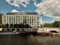 Nowoczesny budynek hotelu Radisson Blu Berlin nad rzeką Sprewą, z widoczną w tle wieżą telewizyjną.