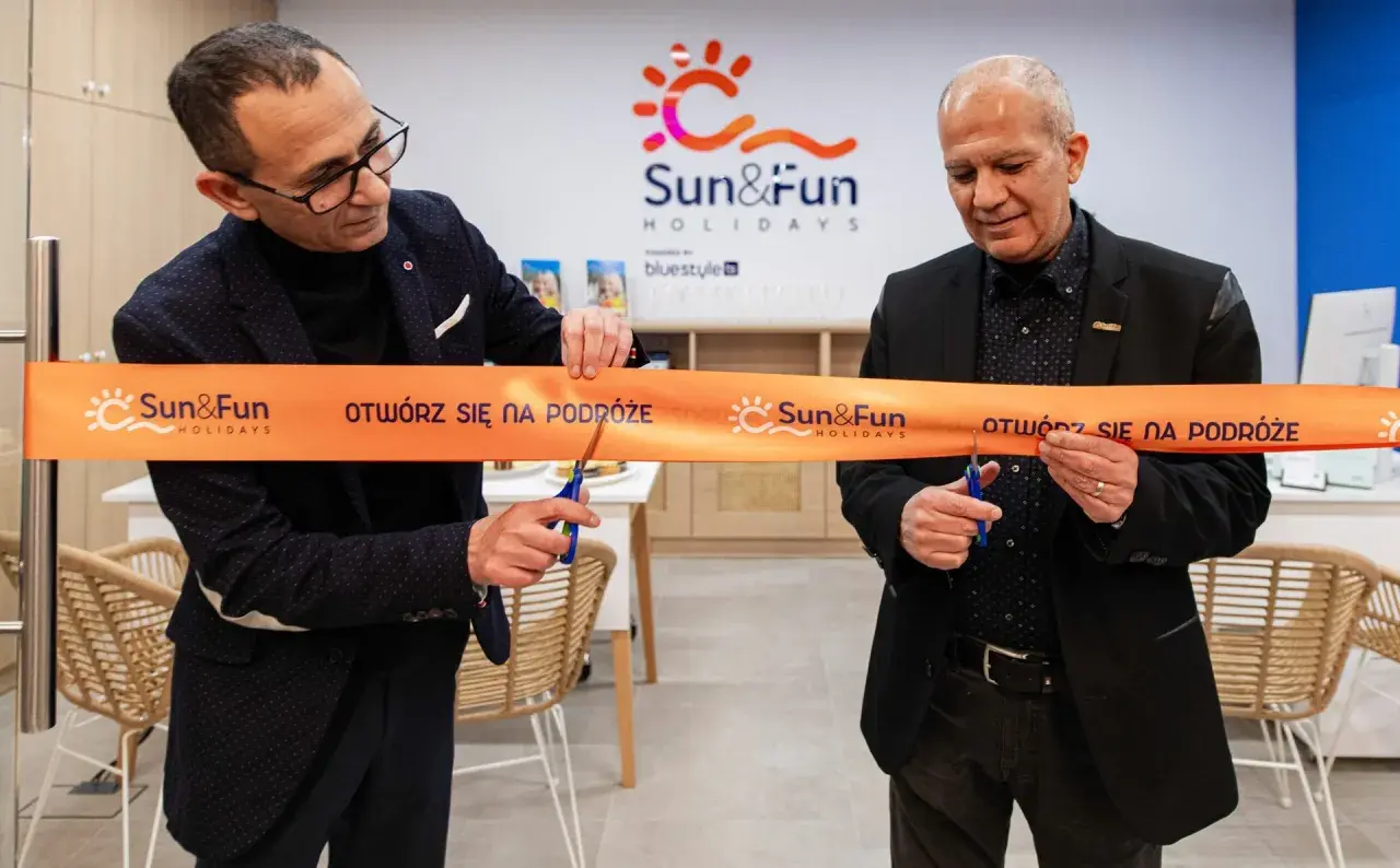 Uroczyste otwarcie biura Sun&Fun Holidays. Dwóch mężczyzn przecina pomarańczową wstęgę z logo firmy, symbolizując nowy początek i zapraszając na podróże.
