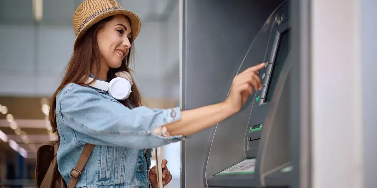 Kobieta w słomkowym kapeluszu i dżinsowej kurtce korzysta z bankomatu.