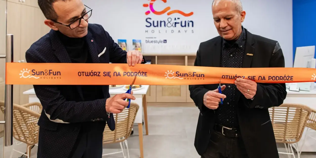 Uroczyste otwarcie biura Sun&Fun Holidays. Dwóch mężczyzn przecina pomarańczową wstęgę z logo firmy, symbolizując nowy początek i zapraszając na podróże.