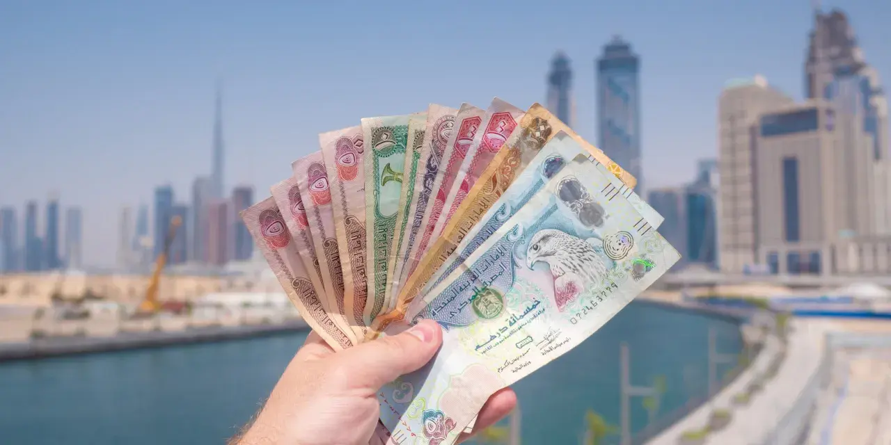 Dłoń trzymająca wachlarz banknotów dirhamów ZEA na tle panoramy Dubaju.