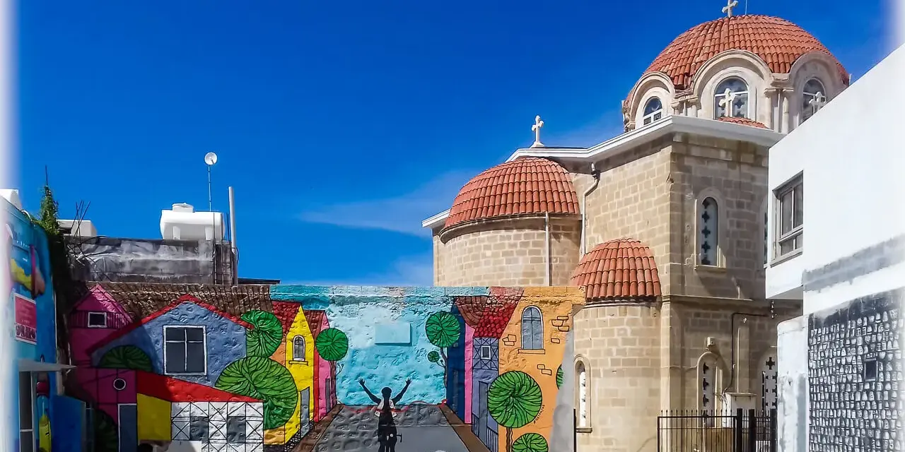 Kolorowy mural z domkami i rowerzystą zdobi ścianę w Pafos, stare miasto. Obok wznosi się kamienna cerkiew z pomarańczowymi kopułami.