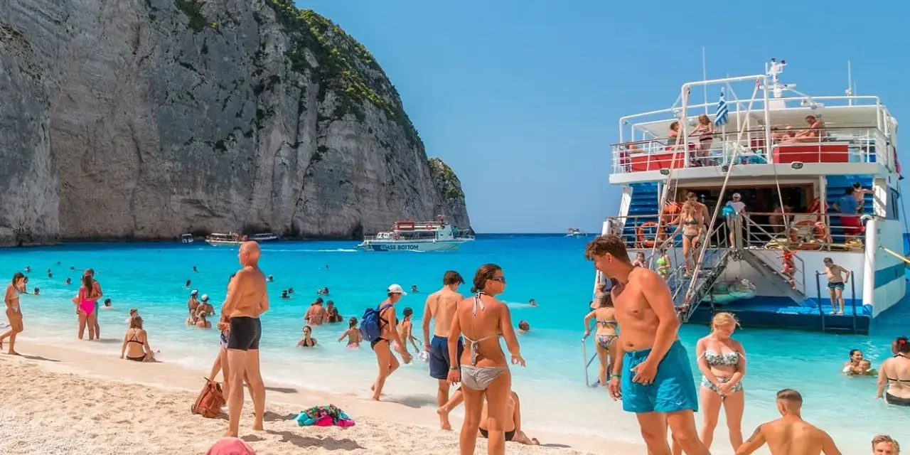 Tui promocje na wakacje: ludzie na plaży Navagio, Zakynthos, Grecja. Turkusowa woda, biały piasek i klify.