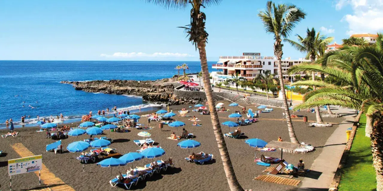 Czarna plaża Playa de la Arena na Teneryfie z turystami, niebieskimi parasolami i palmami.