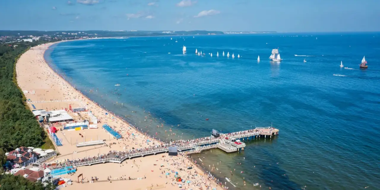 Słoneczna plaża z molo, tłumy ludzi i żaglówki na morzu – idealne miejsce na tanie wycieczki z Gdańska.