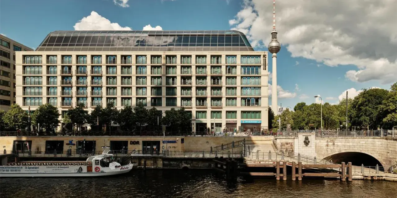 Nowoczesny budynek hotelu Radisson Blu Berlin nad rzeką Sprewą, z widoczną w tle wieżą telewizyjną.