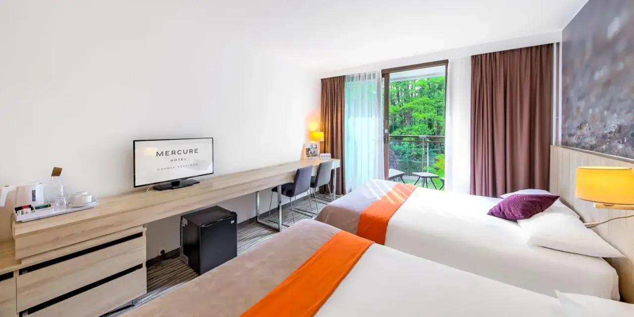Pokój hotelowy z dwoma łóżkami, biurkiem i balkonem z widokiem na zieleń. Hotel Posejdon Gdańsk.