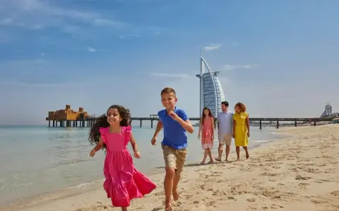 Rodzina cieszy się słonecznym dniem na plaży w Dubaju. Dzieci biegną w stronę morza, a w tle widać słynny hotel Burj Al Arab.