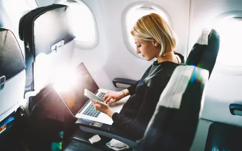 Kobieta pracuje na laptopie i trzyma telefon, zastanawiając się, czy w samolocie można ładować telefon.