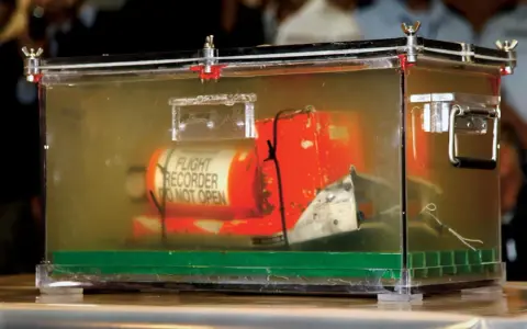 Pomarańczowa skrzynka "czarnej skrzynki" z napisem "FLIGHT RECORDER DO NOT OPEN" w przezroczystej obudowie, dowód w sprawie wypadku lotniczego.