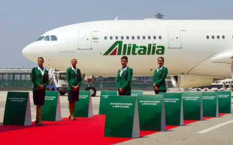 Cztery stewardessy w zielonych uniformach stoją na czerwonym dywanie przed samolotem Alitalia.