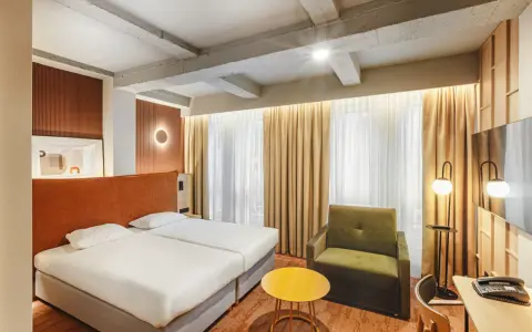Przytulny pokój w hotelu Qubus Wrocław z dwoma łóżkami, zielonym fotelem i stolikiem. Widoczne betonowe belki na suficie.