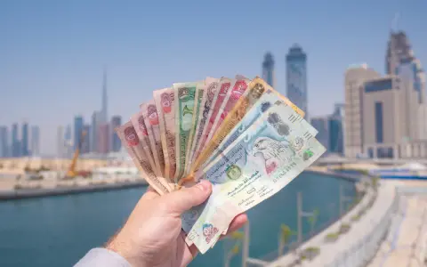 Dłoń trzymająca wachlarz banknotów dirhamów ZEA na tle panoramy Dubaju.