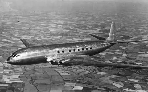 Pierwszy samolot pasażerski, lśniący de Havilland Comet, przelatuje nad patchworkiem pól.