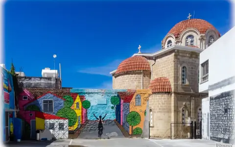 Kolorowy mural z domkami i rowerzystą zdobi ścianę w Pafos, stare miasto. Obok wznosi się kamienna cerkiew z pomarańczowymi kopułami.