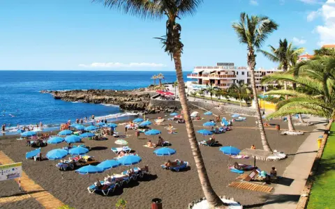 Czarna plaża Playa de la Arena na Teneryfie z turystami, niebieskimi parasolami i palmami.