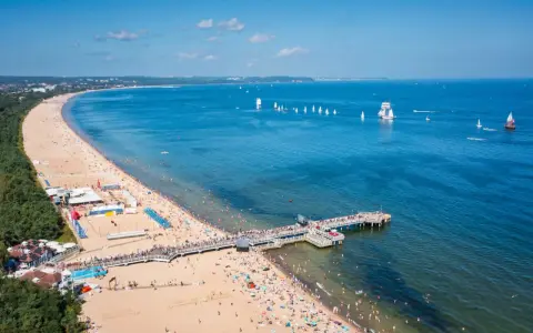 Słoneczna plaża z molo, tłumy ludzi i żaglówki na morzu – idealne miejsce na tanie wycieczki z Gdańska.