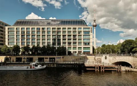 Nowoczesny budynek hotelu Radisson Blu Berlin nad rzeką Sprewą, z widoczną w tle wieżą telewizyjną.
