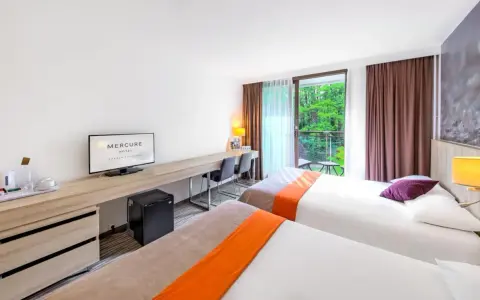 Pokój hotelowy z dwoma łóżkami, biurkiem i balkonem z widokiem na zieleń. Hotel Posejdon Gdańsk.