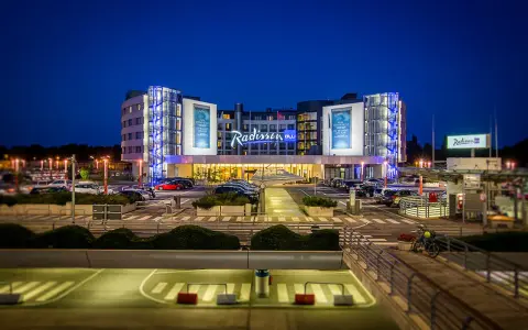 Nowoczesny hotel Radisson Blu Hamburg Airport nocą, z podświetlanym logo i parkingiem pełnym samochodów.