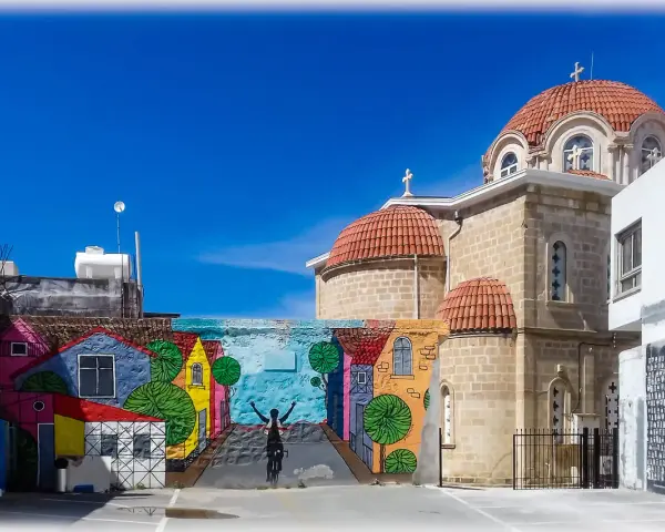 Kolorowy mural z domkami i rowerzystą zdobi ścianę w Pafos, stare miasto. Obok wznosi się kamienna cerkiew z pomarańczowymi kopułami.