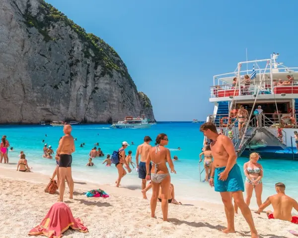 Tui promocje na wakacje: ludzie na plaży Navagio, Zakynthos, Grecja. Turkusowa woda, biały piasek i klify.