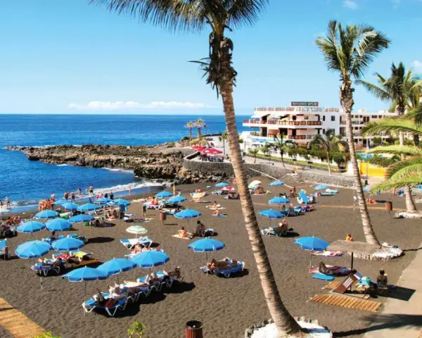 Czarna plaża Playa de la Arena na Teneryfie z turystami, niebieskimi parasolami i palmami.
