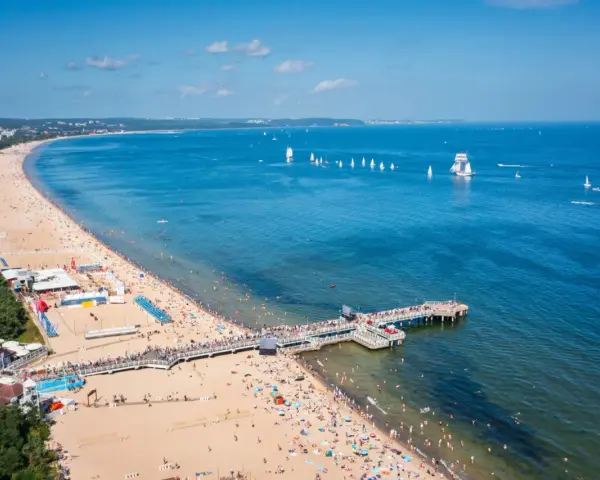 Słoneczna plaża z molo, tłumy ludzi i żaglówki na morzu – idealne miejsce na tanie wycieczki z Gdańska.