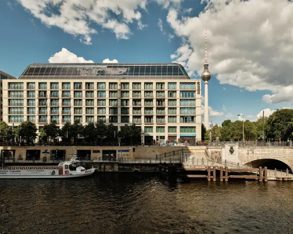 Nowoczesny budynek hotelu Radisson Blu Berlin nad rzeką Sprewą, z widoczną w tle wieżą telewizyjną.