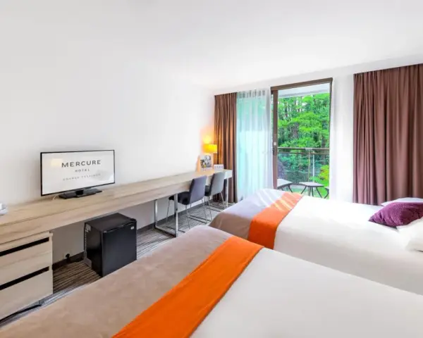 Pokój hotelowy z dwoma łóżkami, biurkiem i balkonem z widokiem na zieleń. Hotel Posejdon Gdańsk.