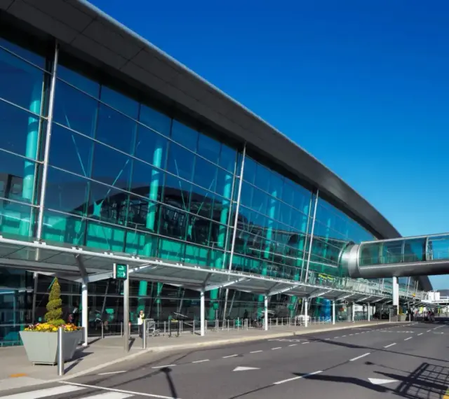 Nowoczesny budynek z przeszkloną fasadą, widoczny jest pas startowy i kładka dla pieszych. To wejście na dublin lotnisko.
