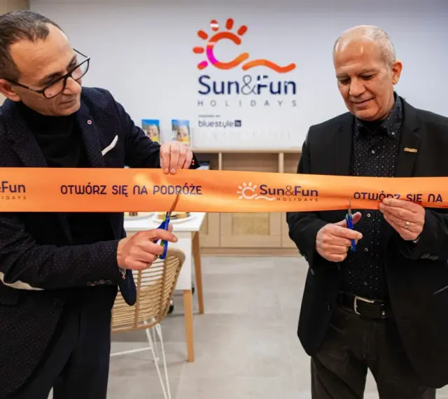 Uroczyste otwarcie biura Sun&Fun Holidays. Dwóch mężczyzn przecina pomarańczową wstęgę z logo firmy, symbolizując nowy początek i zapraszając na podróże.