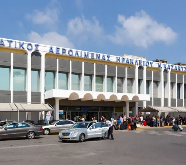 Lotnisko na Krecie, Heraklion N. Kazantzakis. Ludzie z bagażami czekają na transport przed nowoczesnym budynkiem terminalu.
