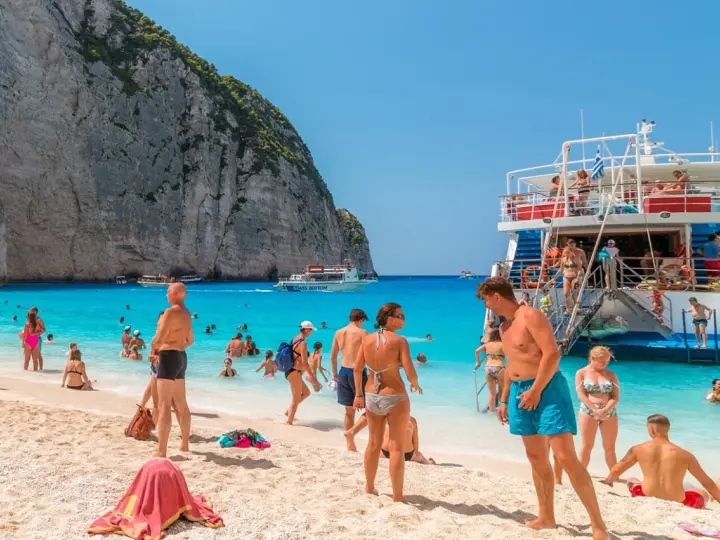 Tui promocje na wakacje: ludzie na plaży Navagio, Zakynthos, Grecja. Turkusowa woda, biały piasek i klify.