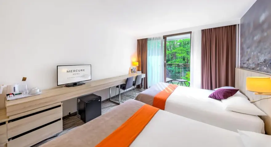Pokój hotelowy z dwoma łóżkami, biurkiem i balkonem z widokiem na zieleń. Hotel Posejdon Gdańsk.