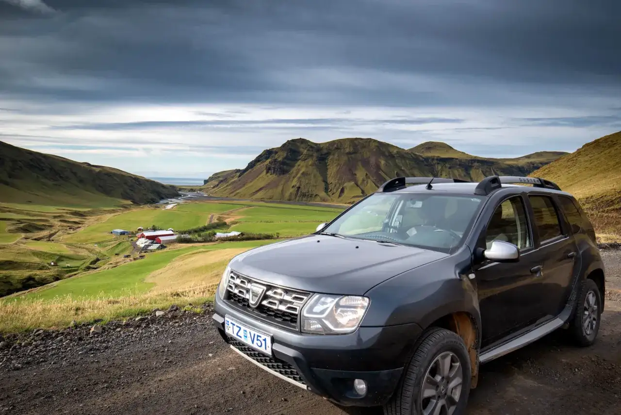 Szara Dacia Duster na drodze z widokiem na dolinę z farmą i górami. Islandia ceny paliwa i noclegów mogą zaskoczyć.