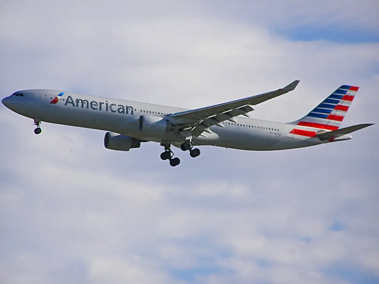 Airbus A330-300 linii American Airlines z wysuniętym podwoziem zbliża się do lądowania na tle błękitnego nieba z chmurami.