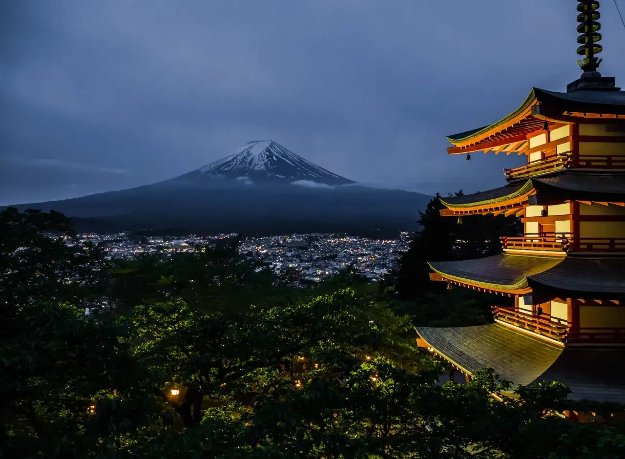 Wieczorem, na tle oświetlonego miasta, widać najwyższy szczyt Japonii – ośnieżony Fudżi. Obok wznosi się wielopiętrowa pagoda.