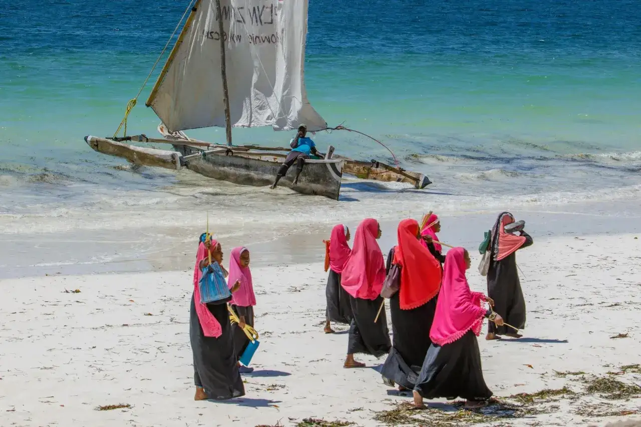 Wycieczka na Zanzibar: grupa kobiet w kolorowych hidżabach spaceruje po plaży, w tle tradycyjna łódź z żaglem.