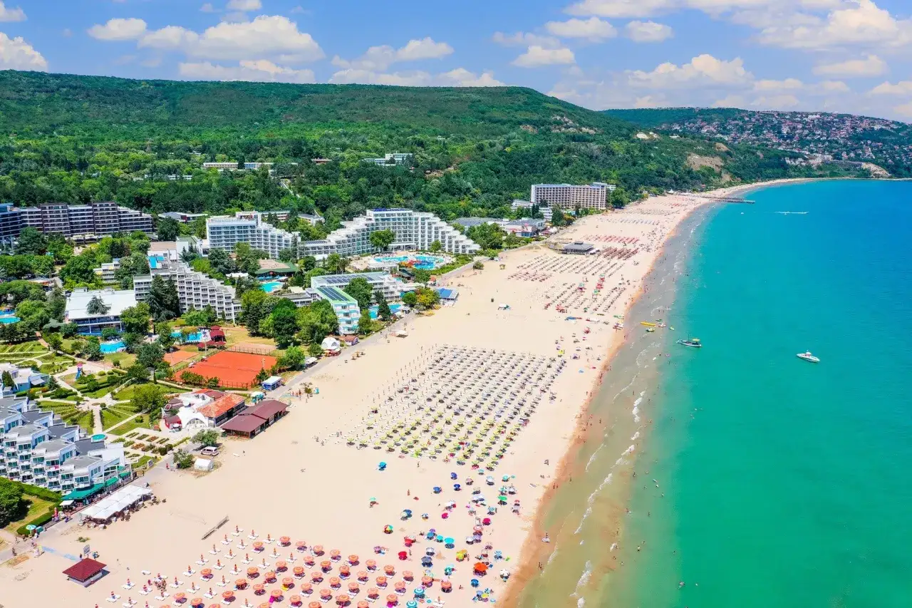 Słoneczna plaża w Bułgarii, pełna turystów i leżaków. W tle hotele i zielone wzgórza. Ceny wakacji w Bułgarii są atrakcyjne.