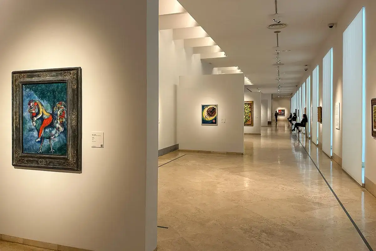 Wnętrze muzeum w Madrycie. Na ścianie wisi obraz z kogutem i postacią. W korytarzu widać inne dzieła i siedzących widzów.