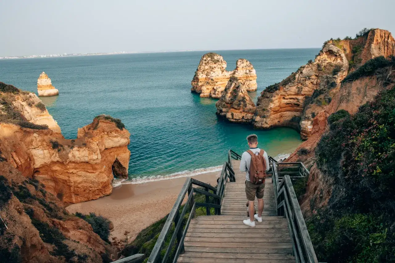 Mężczyzna z plecakiem schodzi drewnianymi schodami ku plaży. Widok na skaliste wybrzeże i turkusowe morze, idealne na portugalskie wakacje.