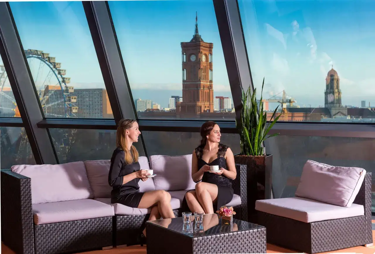 Dwie kobiety piją kawę na sofie z widokiem na Berlin. W tle widać Riesenrad i wieżę ratuszową, charakterystyczne dla Radisson Blu Berlin.