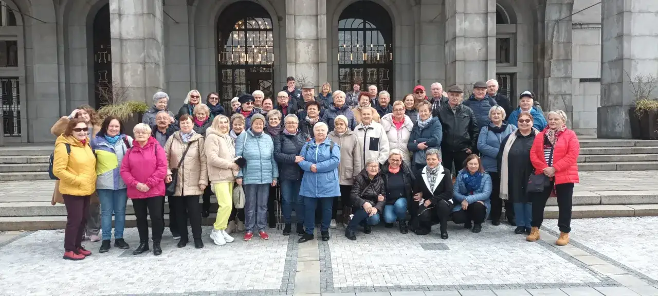 Grupa uśmiechniętych seniorów pozuje przed budynkiem. To z pewnością efekt udanych, ciekawych wycieczek dla seniorów, pełnych radości i wspólnych chwil.